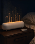 AURALIE Jellyfish Diffuser — horizontal white pill body with chrome legs, multiple translucent jellyfish-shaped mist columns rising from the top grille, warm amber internal LED glow, on a dark walnut nightstand with cream linen bedding at the edge